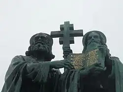 A photograph of statue, which depicts two bearded and hooded men; the one on the left is holding up a cross and the one on the right is holding up a book